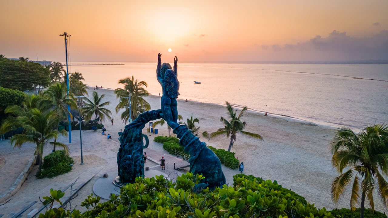 Services Stunning sunset view at Portal Maya sculpture, Playa del Carmen beachside with palm trees and serene sea.